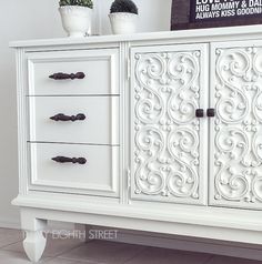 an ornate white cabinet with three drawers and two potted plants sitting on top of it