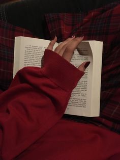 a person with their hand on an open book while laying in bed and wearing a red sweater