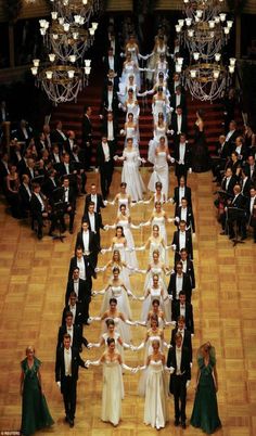 a large group of people in formal wear standing on a wooden floor with chandeliers