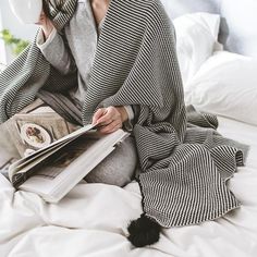 a woman sitting on top of a bed while holding a coffee cup and reading a book