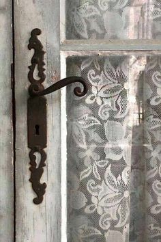 an iron handle on the side of a wooden door with lace curtains behind it and a window in the background