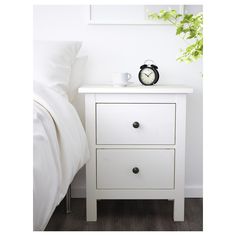 a white nightstand with a clock on it next to a plant and a bed in the background