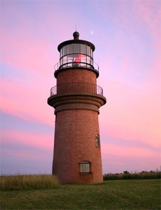 a red and white lighthouse on top of a grassy hill at sunset with pink clouds in the background