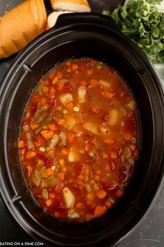 a crock pot filled with vegetable soup next to bread and carrots on a table