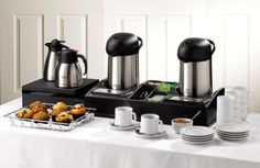 a table topped with plates and cups filled with breakfast foods next to two coffee maker's