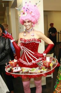 a woman in a pink wig and red dress holding a tray with candies on it