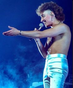 a shirtless man holding a microphone in his right hand and wearing white pants on stage