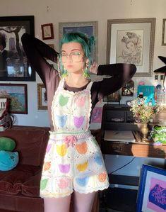 a woman with green hair and glasses standing in front of a couch wearing colorful clothing