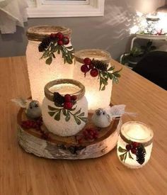 three mason jars decorated with holly and pine cones are sitting on a wooden table next to candles