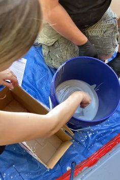 two people are working on something in a blue bucket with water inside and one person is holding a piece of paper