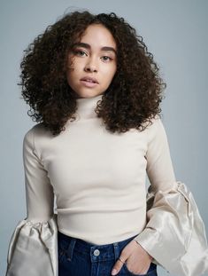 a woman with curly hair wearing a white top and blue jeans is posing for the camera