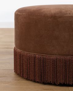 a brown round ottoman with fringe trim on the top and bottom, sitting on a hard wood floor