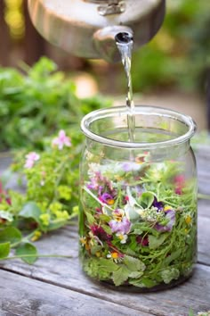 a glass jar filled with flowers and water on top of a wooden table next to plants