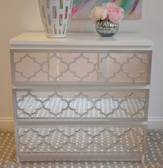 a white dresser with three drawers and a vase on top of it next to a painting