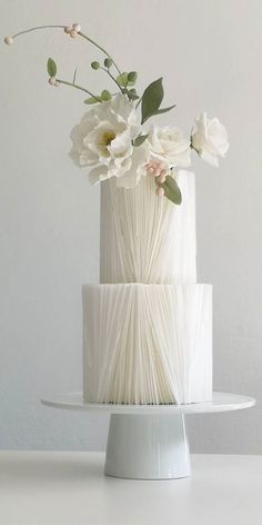 a three tiered white cake with flowers in the middle on a stand against a wall