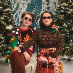 two people sitting next to each other in front of a christmas tree wearing ugly sweaters