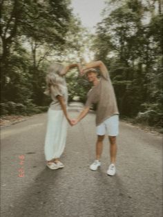 two people holding hands while standing in the middle of a road with trees behind them