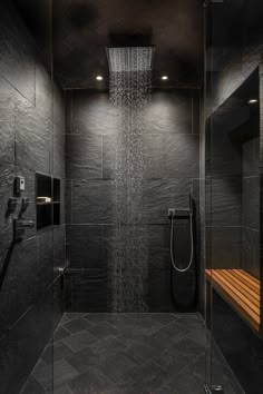 a shower head in the middle of a black tiled bathroom with wooden bench under it