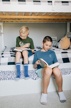 two boys sitting on the bottom bunk of a bed reading books and looking at each other