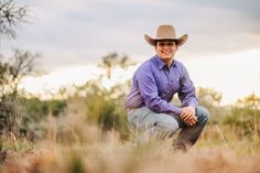 a man wearing a cowboy hat sitting in the grass with his hands on his knees