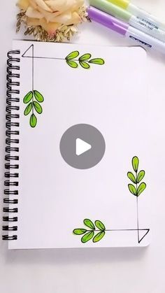 a spiral notebook with green leaves on it next to two markers and a flower arrangement