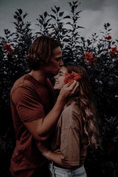 a man and woman standing next to each other in front of some red flowers with their faces close together