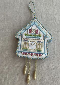 a cross stitch house ornament hanging on a gray fabric background with gold chain