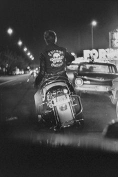 black and white photograph of man on motorcycle in street at night with car passing by