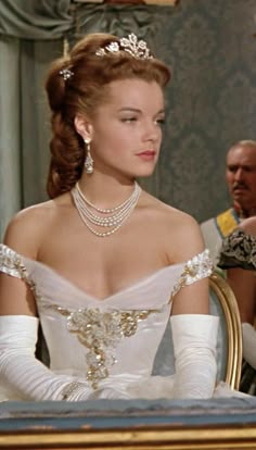 a woman wearing a tiara and pearls sits at a table in front of other people