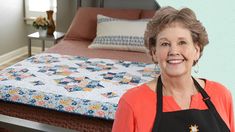 a woman in an apron standing next to a bed with a quilt on the headboard