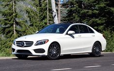 a white mercedes benz is parked on the side of the road in front of some trees