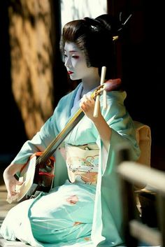 a geisha doll sitting on the ground with a guitar in her hand and wearing a blue kimono