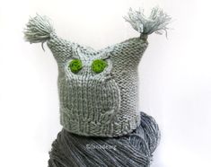 a gray knitted owl hat with green eyes on top of a ball of yarn