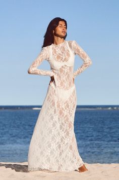 a woman is standing on the beach wearing a white lace dress with long sleeves and an open back