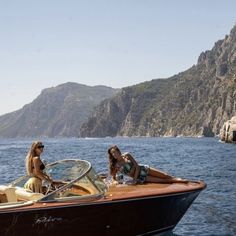 two women are sitting on the back of a boat in the water near some mountains