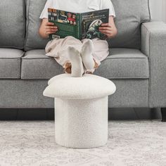 a young boy reading a book while sitting on top of a white stool in front of a gray couch