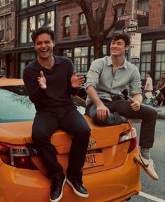 two men sitting on top of an orange car in the middle of a city street