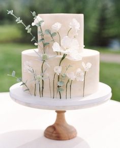 a three tiered cake with white flowers and greenery on the top is sitting on a table outside