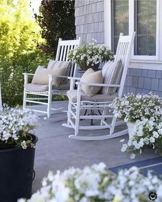 two white rocking chairs sitting on top of a porch next to flowers and bushes in front of a house