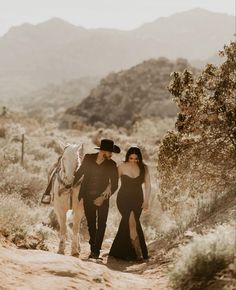 a man and woman walking down a dirt road with a horse in the desert behind them