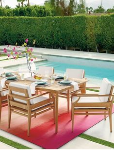 an outdoor dining table with white chairs and pink rug next to swimming pool in backyard