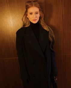 a woman standing in front of a wooden wall wearing a black coat and turtle neck sweater