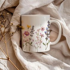 a coffee cup with flowers painted on it sitting on a white blanket next to dried plants