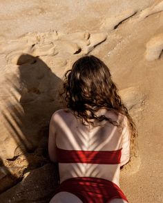 a woman in a red and white striped bathing suit sitting on the sand with her back to the camera