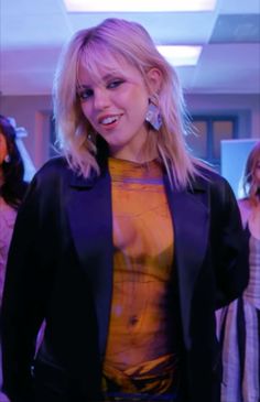 a woman with blonde hair wearing a black jacket and yellow top standing in front of other women