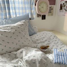 an unmade bed with blue and white sheets, pillows and a clock on the wall