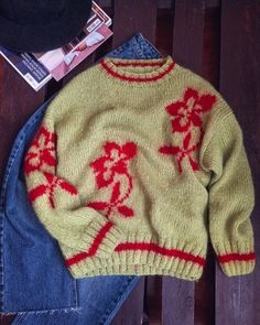 a yellow sweater with red flowers is sitting on a wooden bench next to jeans and a hat