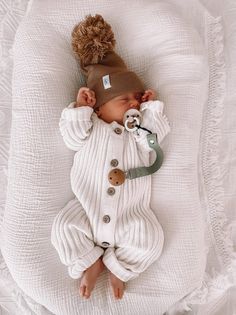 a baby is laying down wearing a white outfit and a brown knitted hat with pom - poms