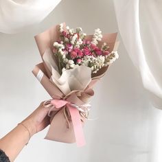 a person holding a bouquet of flowers in their hand with white curtains behind them and pink ribbon around the edges