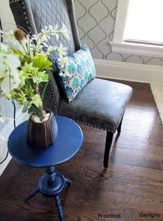 a blue table with a vase on it next to a gray chair and flower arrangement